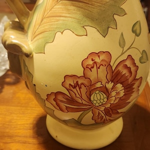 May include: A cream-colored ceramic pitcher with a floral design. The pitcher has a wide mouth and a handle. The floral design features red, green, and brown flowers and leaves.