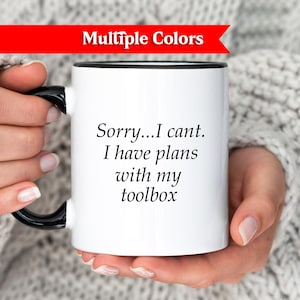 Puede incluir: Taza de cerámica blanca con asa y borde negros, con el texto "Sorry...I cant. I have plans with my toolbox." Una pancarta roja en la parte superior dice "Multiple Colors."