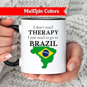 May include: White ceramic mug with a black handle and rim, featuring the text "I don't need THERAPY I just need to go to BRAZIL" and a green and yellow map of Brazil. The mug is held by a person wearing a white sweater.