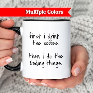 Könnte beinhalten: Eine weiße Kaffeetasse mit schwarzem Griff und Rand, mit dem Text "first i drink the coffee. then i do the Coding things." Die Tasse wird von einer Person gehalten, die einen grauen Pullover trägt. Ein rotes Banner oben zeigt "Multiple Colors."