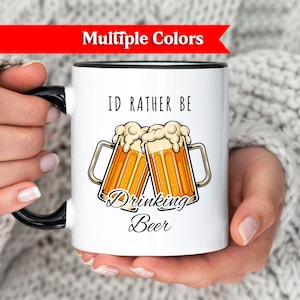 Può includere: Tazza in ceramica bianca con manico e bordo neri, con la scritta "ID RATHER BE Drinking Beer" e un'illustrazione di due boccali di birra. La tazza è tenuta da una persona che indossa un maglione bianco.