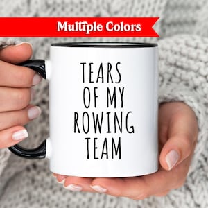 May include: White ceramic mug with a black handle and rim, featuring the text "TEARS OF MY ROWING TEAM" in black. A red banner at the top reads "Multiple Colors". The mug is held by a person wearing a grey sweater.
