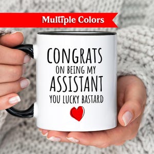 May include: White ceramic mug with black handle and rim, featuring the text "CONGRATS ON BEING MY ASSISTANT YOU LUCKY BASTARD" and a red heart. The mug is held by a person wearing a white sweater.