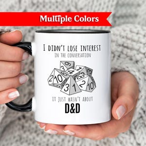 Peut inclure: Mug en céramique blanche avec une anse et un bord noirs, avec le texte "I DIDN'T LOSE INTEREST IN THE CONVERSATION IT JUST WASN'T ABOUT D&D" et des graphiques de dés. Le mug est tenu par une personne portant un pull blanc.
