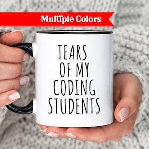Könnte beinhalten: Weiße Keramiktasse mit schwarzem Henkel und Rand, mit dem Text "TEARS OF MY CODING STUDENTS" in Schwarz. Die Tasse wird von einer Person gehalten, die einen grauen Pullover trägt. Ein rotes Banner oben zeigt "Multiple Colors".
