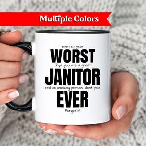 Peut inclure: Mug à café blanc avec anse et bord noirs, avec le texte "even on your WORST days you are a great JANITOR and an amazing person, don't you EVER Forget it!". Une bannière rouge en haut indique "Multiple Colors".
