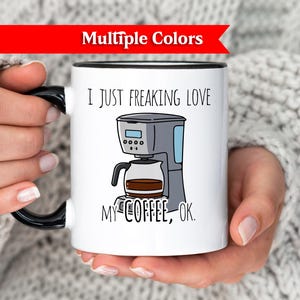 Op de afbeelding: Witte koffiemok met een zwarte handgreep en rand, met een cartoon illustratie van een koffiezetapparaat en de tekst "I JUST FREAKING LOVE MY COFFEE, OK." Een rode banner bovenaan leest "Multiple Colors."