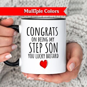May include: White ceramic mug with black handle and rim, featuring the text "CONGRATS ON BEING MY STEP SON YOU LUCKY BASTARD" in black, with a red heart. The mug is held by a person wearing a gray sweater.