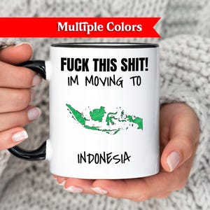 Op de afbeelding: Witte keramische mok met zwarte handgreep en rand, met de tekst "FUCK THIS SHIT! I'M MOVING TO" boven een groene omtrek van Indonesië, met "INDONESIA" eronder. De mok wordt door een persoon vastgehouden.