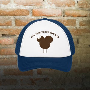 Op de afbeelding: Witte en blauwe truckercap met een bruine Mickey Mouse ijsje-afbeelding en de tekst "It's time to hit the bar".