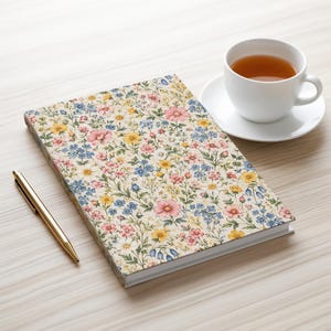 May include: A rectangular notebook featuring a cream-colored cover with a dense, colorful floral pattern of pink, yellow, and blue flowers. A gold-toned ballpoint pen lies to the left, and a white ceramic teacup filled with tea sits nearby.