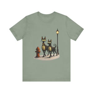 May include: Sage green t-shirt featuring a whimsical illustration of two black cats with glowing eyes, standing near a fire hydrant and a lamppost. The cats have ornate designs on their sides. The design is set against a snowy ground.