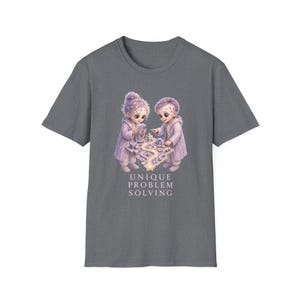 Peut inclure: Un t-shirt gris anthracite à manches courtes orné d'un motif représentant deux enfants en robes violet clair interagissant avec un plateau de jeu lumineux. Sous l'illustration, le texte UNIQUE PROBLEM SOLVING est écrit en lettres capitales blanches.