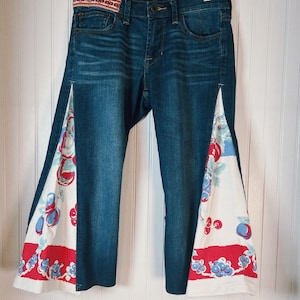 May include: Blue denim jeans with a decorative red and white patterned waistband. The sides of the jeans feature a white fabric panel with a red and blue floral design. The jeans are hanging on a wooden hanger.