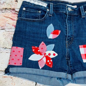 May include: Blue denim shorts with red and white patchwork details. The shorts have a flower design on the front and a white lace waistband.