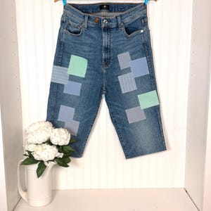 Pode incluir: Jeans capri de ganga azul com remendos de vários tecidos listrados e lisos azuis e verdes. Os jeans estão pendurados em um cabide de madeira contra uma parede branca com painéis. Um jarro branco com flores brancas está em uma prateleira abaixo.
