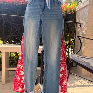 Peut inclure: Jean en denim bleu clair avec une coupe taille haute et des jambes droites. Les coutures latérales extérieures sont ornées de larges bandes verticales en tissu rouge vif, décorées d'un motif floral et fruité blanc, avec des ourlets effilochés.
