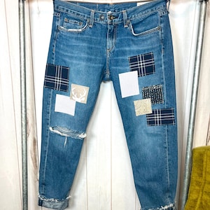May include: Blue denim jeans with patches of various patterns, including plaid, stripes, and a deer silhouette. The jeans have a distressed knee and rolled-up cuffs, hanging on a metal rack.