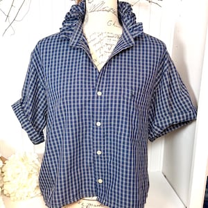 May include: A blue and white checked short-sleeved shirt with a ruffled collar and button-down front. The shirt is displayed on a mannequin. The sleeves are cuffed. The shirt has a small logo on the chest.