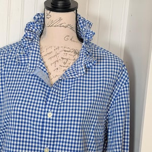 Puede incluir: Camisa a cuadros azul y blanco con cuello con volantes. La camisa de manga larga está abotonada y expuesta en un maniquí. La tela tiene un diseño de cuadros clásico.