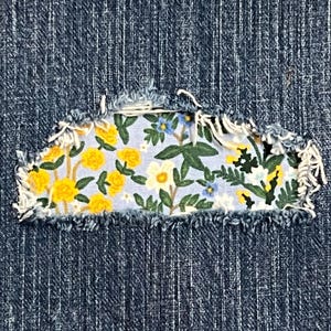 Puede incluir: Un primer plano de una tela vaquera con un parche de estampado floral cosido sobre un agujero. El parche presenta flores amarillas, hojas verdes y un fondo azul claro.