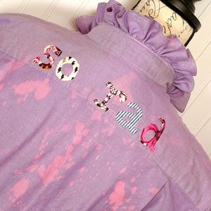 May include: A lavender button-down shirt with pink bleach-splatter design and the embroidered word "SOFIA" across the back. The shirt has a ruffled collar and is displayed on a mannequin.