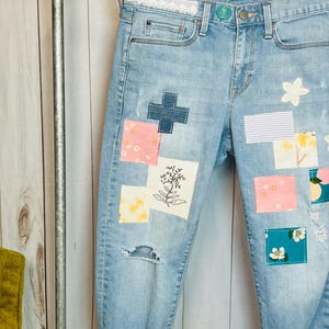 May include: Light blue denim jeans with various fabric patches sewn on. The patches feature floral designs, stripes, and a cross.