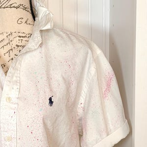Puede incluir: Camisa blanca de manga corta con un diseño de pintura salpicada en rosa y turquesa. La camisa tiene una botonadura frontal, un cuello clásico y un pequeño logotipo azul marino en el pecho. Las mangas están dobladas.