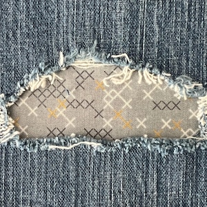 May include: Close-up of a denim patch with frayed edges, sewn onto a blue denim background. The patch features a gray fabric with a repeating pattern of white, black, and yellow cross shapes. The patch is a unique design element.