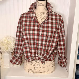 May include: A long-sleeved button-down shirt featuring a red, white, and dark green plaid pattern. The shirt has a ruffled collar, a front button closure, and long sleeves with folded cuffs. The hem is tied in a knot at the front waist area.