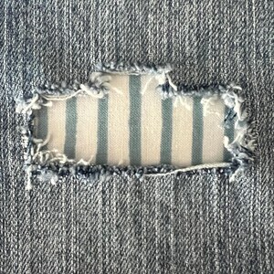 May include: Close-up of a denim fabric with a rectangular patch. The patch is made of a blue and white striped fabric, and the edges of the denim are frayed.