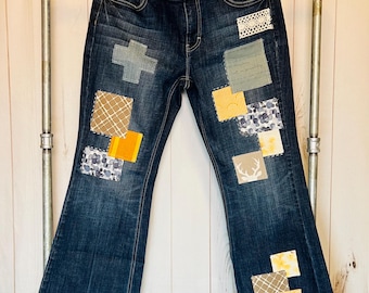 May include: Blue denim jeans with patchwork details in shades of brown, yellow, and white. The patches include geometric shapes, floral patterns, and a deer silhouette.