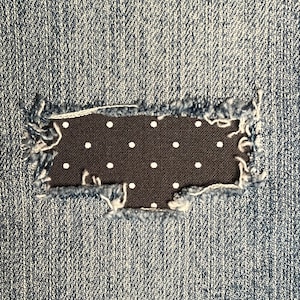 May include: Close-up of a denim fabric with a rectangular patch. The patch is a dark fabric with white polka dots, framed by the frayed edges of the denim. The denim has a blue wash.