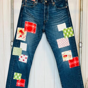 May include: Blue denim jeans with patchwork details. The patches are made of various fabrics with floral, polka dot, and plaid patterns in red, white, and green.