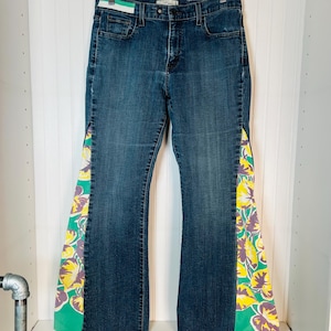 May include: Dark wash denim jeans with flared legs. The sides of the jeans feature a colourful floral pattern in yellow, purple, and green. The jeans are hanging on a wooden hanger.