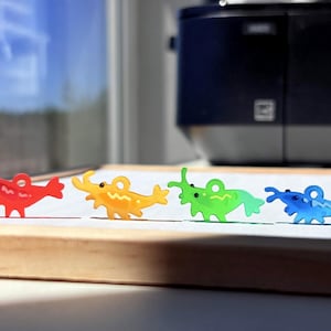 Puede incluir: Cuatro dijes de acrílico translúcido con forma de camarón, cada uno con un pequeño lazo circular en la parte superior. Los dijes están dispuestos en fila y son de color rojo, amarillo, verde y azul, con un ojo y detalles grabados simples.