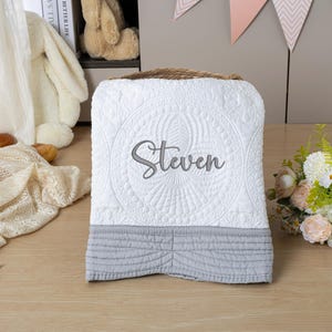 May include: White quilted baby blanket with the name "Steven" embroidered in gray. The blanket has a gray bottom border and a decorative stitched pattern. A woven basket handle is visible at the top.