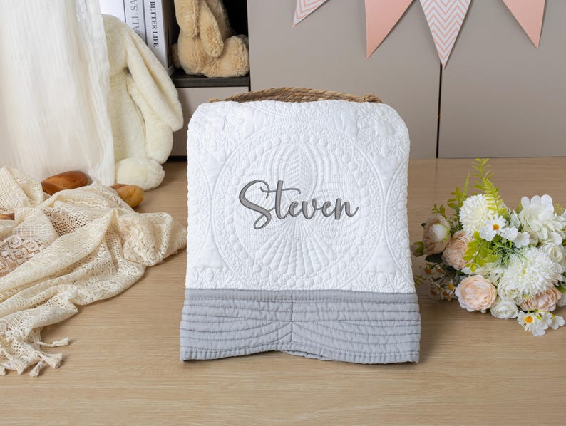May include: White quilted baby blanket with the name "Steven" embroidered in gray. The blanket has a gray bottom border and a decorative stitched pattern. A woven basket handle is visible at the top.