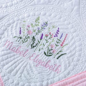 May include: White quilted blanket with a pink border. The blanket features an embroidered floral design in shades of green, pink, and purple, with the name "Mabel Elizabeth" embroidered in pink script. The quilting pattern includes intricate designs.