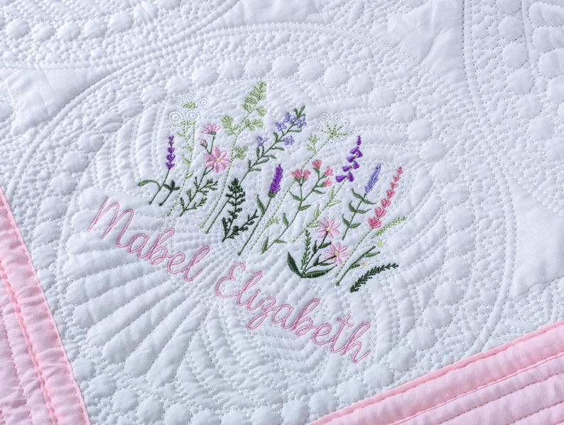 May include: White quilted blanket with a pink border. The blanket features an embroidered floral design in shades of green, pink, and purple, with the name "Mabel Elizabeth" embroidered in pink script. The quilting pattern includes intricate designs.