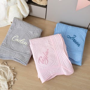 May include: Three embroidered baby blankets in gray, pink, and blue. Each blanket features a unique name: "Caden", "Taylor", and "Jason". The blankets have a quilted design with a central circular pattern.