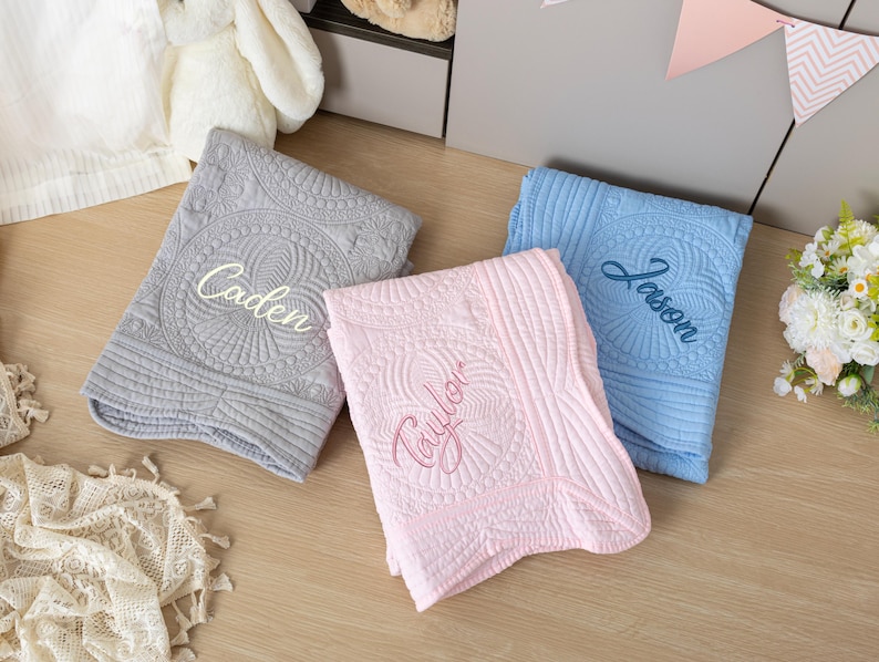 May include: Three embroidered baby blankets in gray, pink, and blue. Each blanket features a unique name: "Caden", "Taylor", and "Jason". The blankets have a quilted design with a central circular pattern.