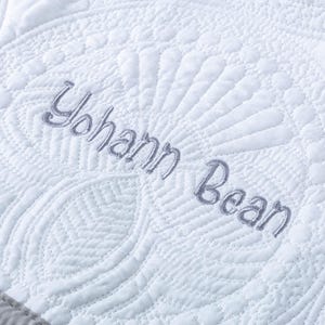 May include: Close-up of a white quilted blanket with intricate patterns and the embroidered name "Yohann Bean" in gray thread. The blanket features a detailed, textured design with a decorative border.