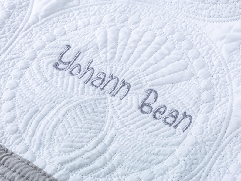 May include: Close-up of a white quilted blanket with intricate patterns and the embroidered name "Yohann Bean" in gray thread. The blanket features a detailed, textured design with a decorative border.