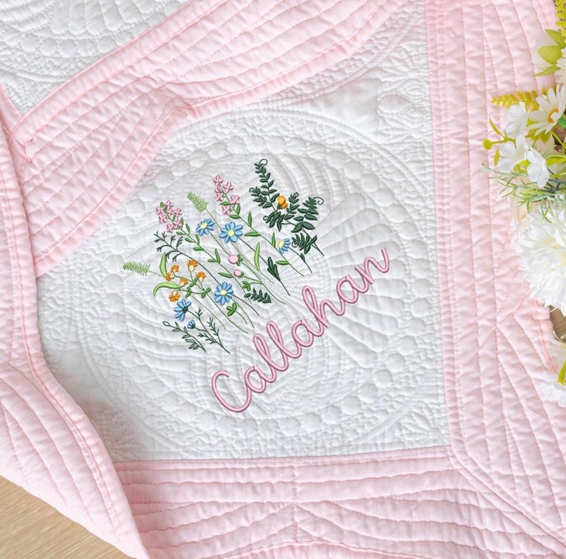 May include: A white and pink quilted blanket with floral embroidery and the name "Callahan" in pink script. The blanket features a detailed floral design in the center, with pink borders and stitching. The overall aesthetic is soft and delicate.