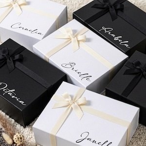 May include: A collection of rectangular gift boxes in black and white, each featuring a satin ribbon bow and a personalized name in script font. The names Cornelia, Arabella, Brielle, Octavia, Paloma, and Janelle are printed on the lids.