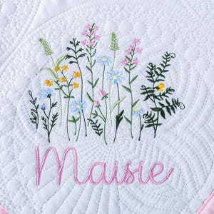 May include: White quilted blanket with embroidered wildflowers in yellow, blue, and pink. The name "Maisie" is embroidered in pink cursive script below the flowers. A pink border frames the blanket.