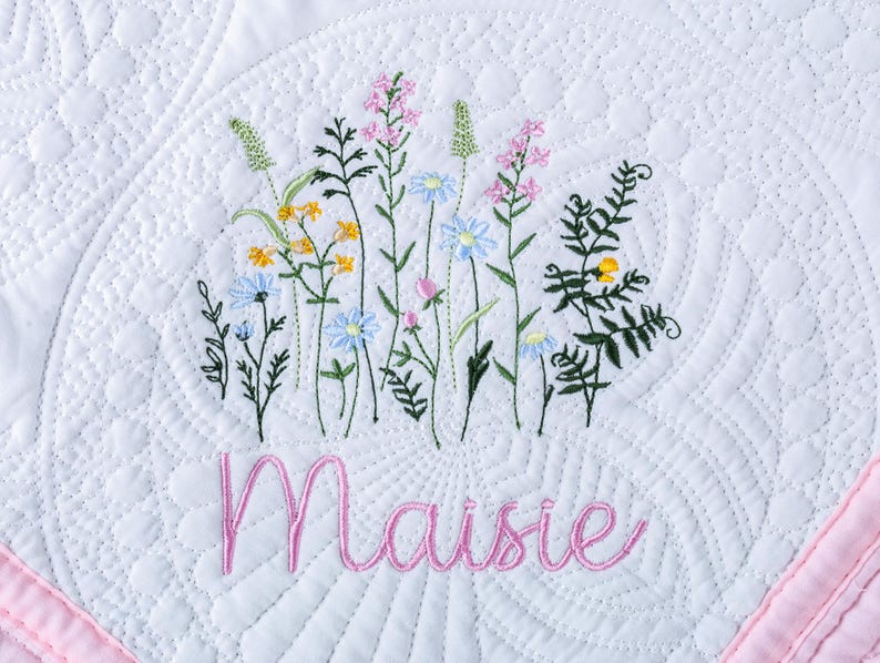 May include: White quilted blanket with embroidered wildflowers in yellow, blue, and pink. The name "Maisie" is embroidered in pink cursive script below the flowers. A pink border frames the blanket.