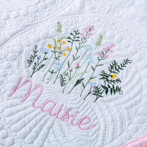 May include: White quilted blanket with a pink border and embroidered floral design. The word "Maisie" is embroidered in pink script below the flowers. The quilt features a detailed, textured pattern.