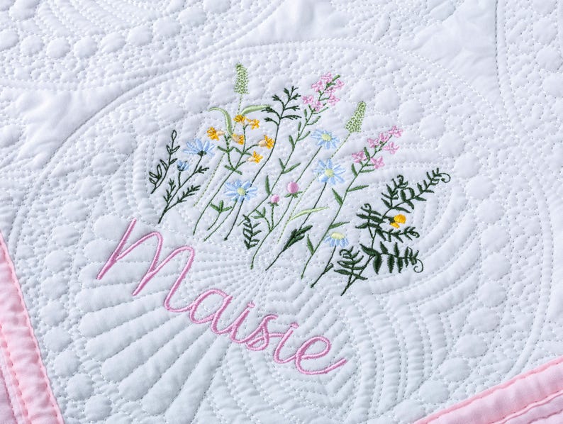 May include: White quilted blanket with a pink border and embroidered floral design. The word "Maisie" is embroidered in pink script below the flowers. The quilt features a detailed, textured pattern.